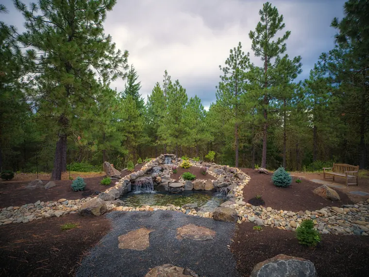 Finished pond with stacked-stone waterfalls, a flagstone path and new planted beds under tall pines at Tumble Creek